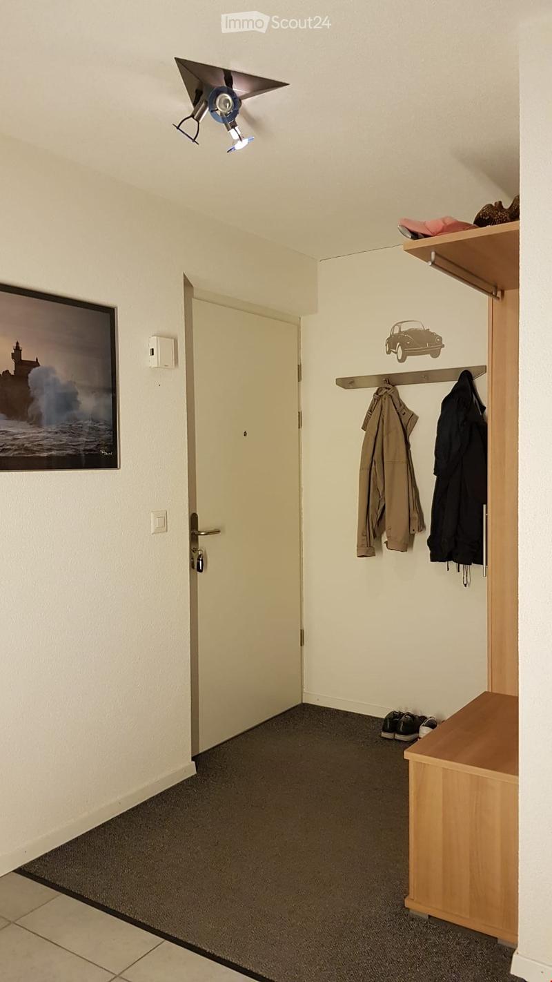 A small, well-lit hallway with a grey carpeted floor, a wooden bench, coat hooks, and a door leading to another room.