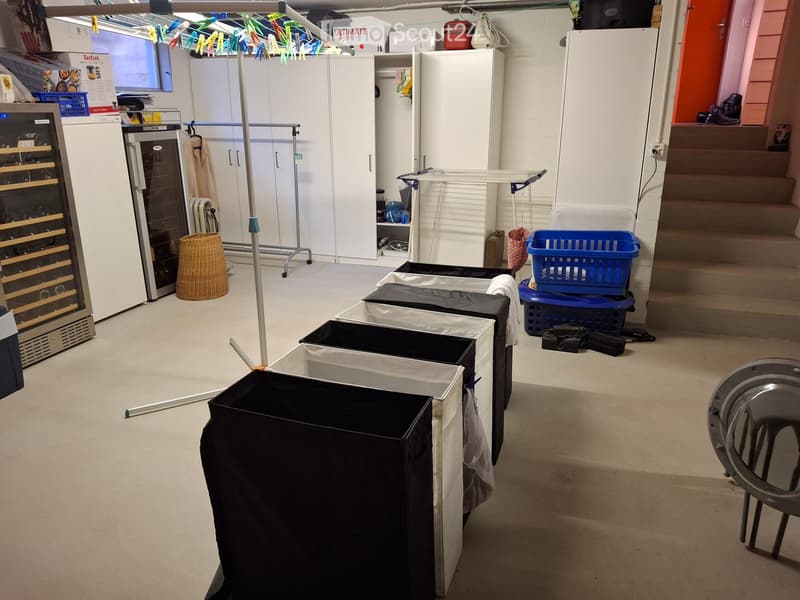 A partially furnished room with a wine cooler, white wardrobes, a laundry drying rack, a wicker basket, a metal chair, and several covered storage bins.