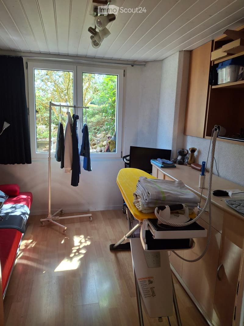 Well-lit room with wooden flooring, a bed, an ironing board, and a cabinet. The room has large windows with a view of greenery outside.