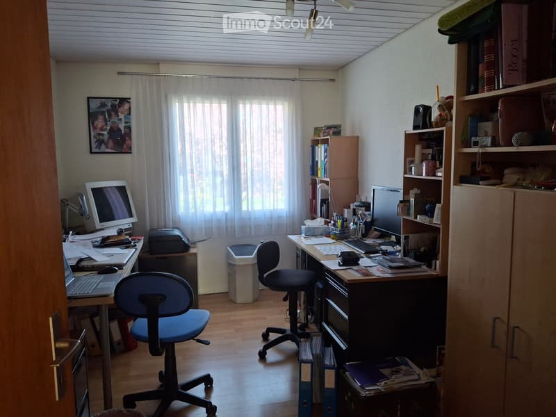 A well-lit office space with two desks, two chairs, a laptop, a screen, a keyboard, a printer, a bookshelf, a speaker, a lamp, and a picture frame on the wall.