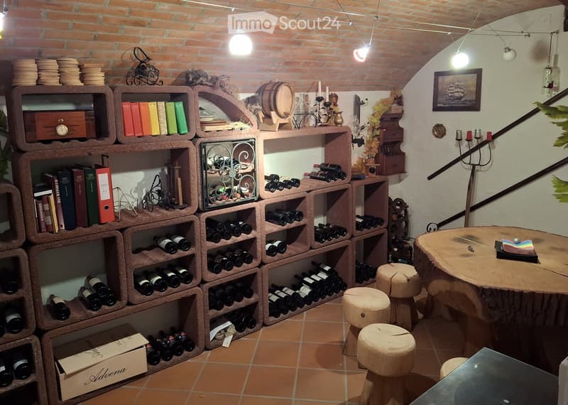 Wine cellar with brick walls, wooden shelves filled with wine bottles, a round wooden table, wooden stools, books, candles, and decorative items.