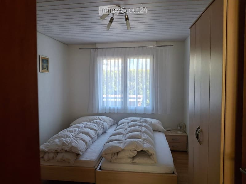 Cozy bedroom with a single bed, nightstand, fan, and picture frame on the wall. The room has white walls, a slatted ceiling, and large windows with sheer curtains.