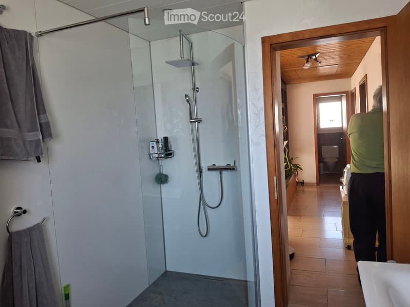 Modern bathroom with a glass-enclosed shower, towel rack, and a visible toilet in the adjacent room.