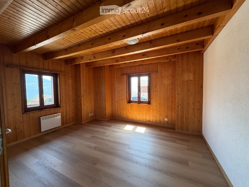 Empty room with wooden walls, wooden ceiling beams, wooden flooring, and two windows offering a view. A white radiator is mounted on one wall.