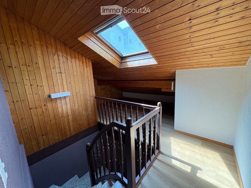 Wooden staircase with a dark railing and a skylight above, leading to an attic space with wooden walls and ceiling.