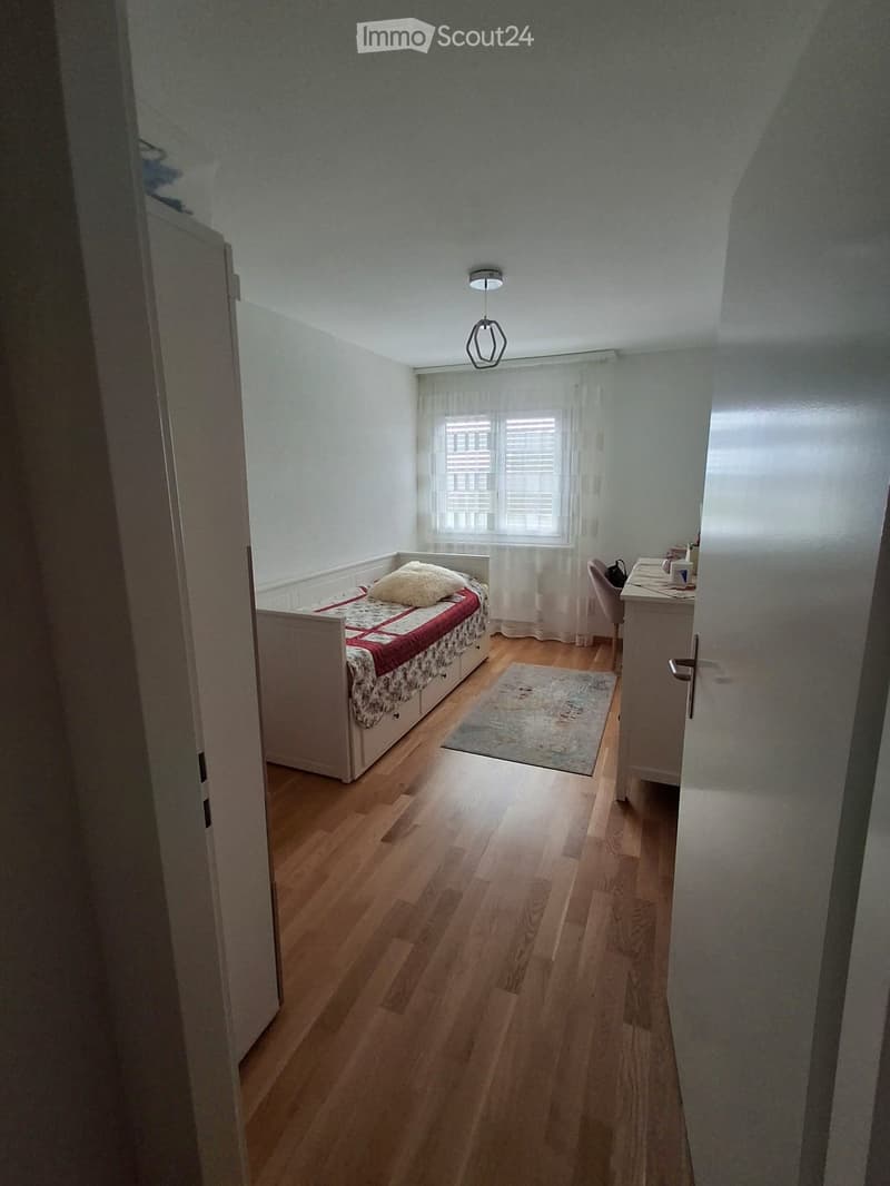 Cozy bedroom with wooden flooring, a single bed with a red and white quilt, a small table with a chair, a rug, and a window with white curtains.