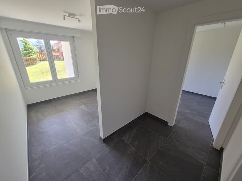 Empty room with dark tiled floor and white walls, featuring a large window with a view of a garden and a door leading to another room.