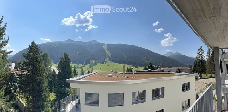 Modern white building with a flat roof and multiple windows, overlooking a scenic mountain landscape with ski slopes and forested areas.