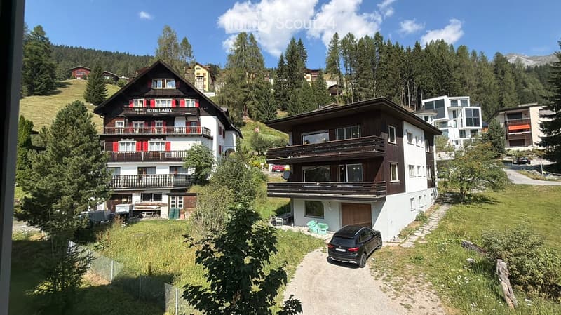 A scenic view of a multi-story wooden house with balconies and red shutters, surrounded by lush greenery and other houses in a mountainous area.