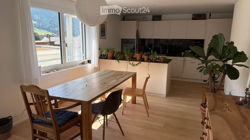 Bright, modern kitchen and dining area with wooden flooring, large windows offering a scenic mountain view, a long wooden dining table with chairs, and a sleek kitchen with cabinets, an oven, and a sink.