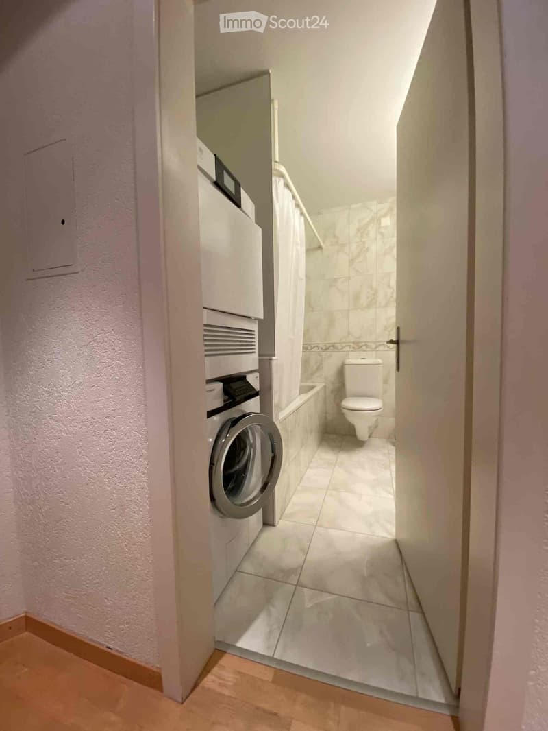 Modern bathroom with a washing machine, toilet, and bathtub, featuring white tiled walls and floor.