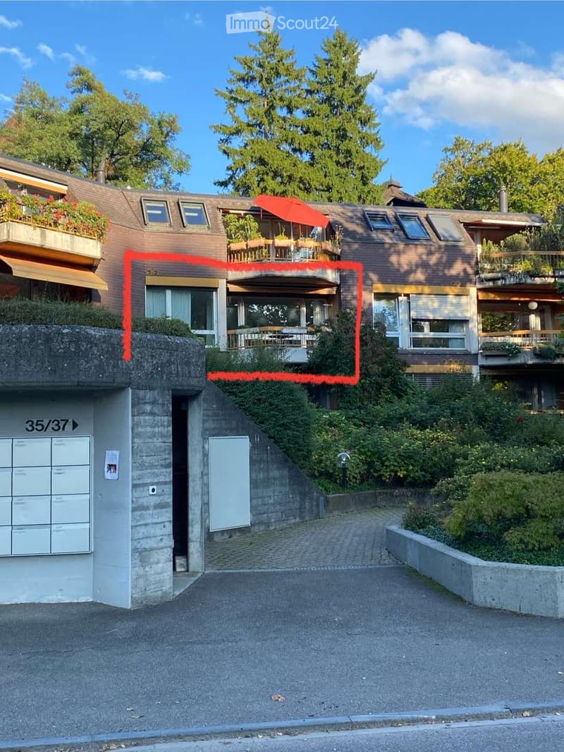 Modern multi-story house with balconies, raised ground floor, and garage. Features large windows, a red umbrella on the balcony, and surrounding greenery.