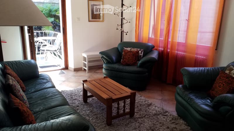 Living room with green leather sofa, armchair, coffee table, and colorful pillows. Large windows with orange curtains and access to a balcony.