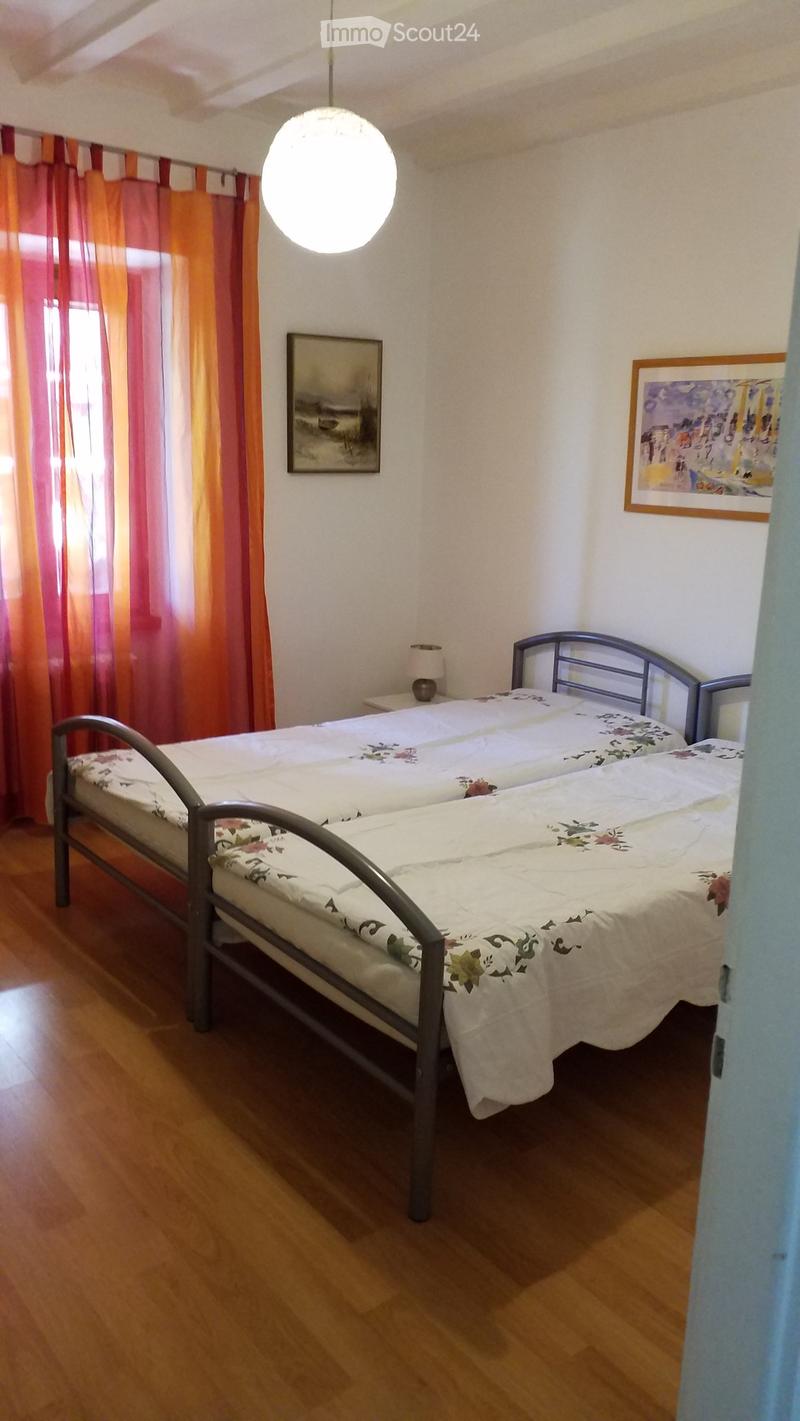 Cozy bedroom with two single beds, wooden flooring, and colorful curtains. Features a nightstand with a lamp and two picture frames on the wall.