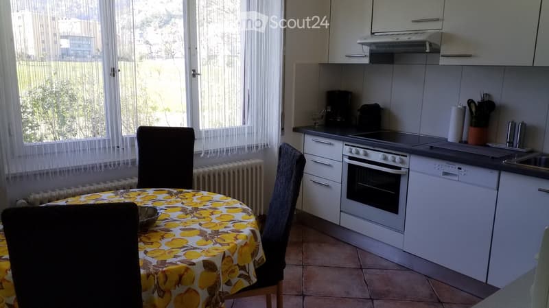 A modern kitchen with white cabinets, a built-in oven, dishwasher, and a dining area with a table and chairs. The room features large windows with white curtains and a radiator below them.