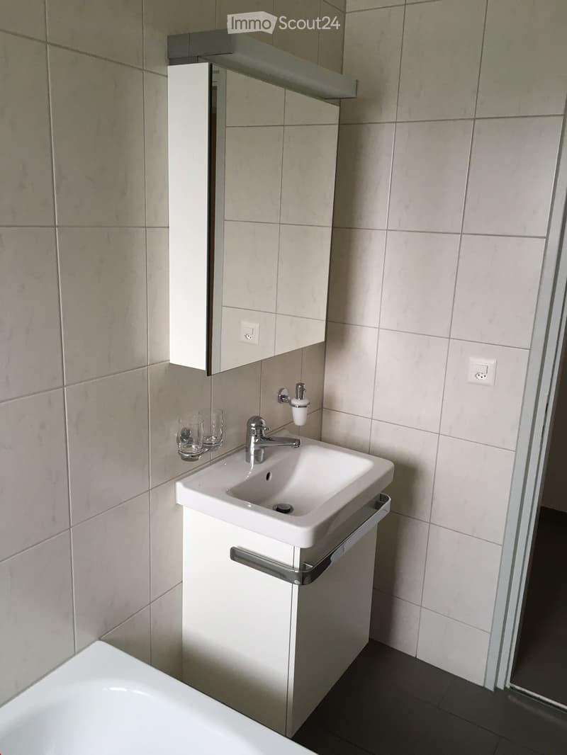 A small, clean bathroom with a white bathtub, sink, and tiled walls. It includes a white wall-mounted cabinet with a mirror and a built-in soap dispenser. There are no visible features such as a balcony, raised ground floor, garage, parking place, swimmin