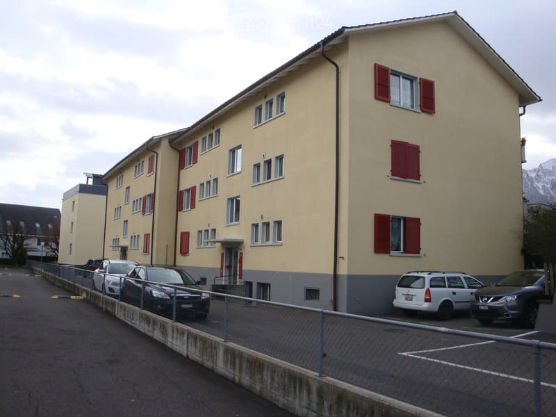 Three story building with red shutters, cars parked in front of the building, a chain link fence and a grey concrete barrier