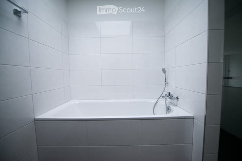 Modern bathroom with white tiled walls and floor, featuring a bathtub with a handheld showerhead and faucet.