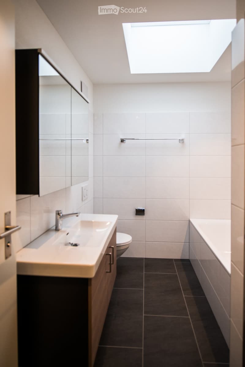 Modern bathroom with white tiles, a large skylight, a sink with a modern faucet, a toilet, and a bathtub.