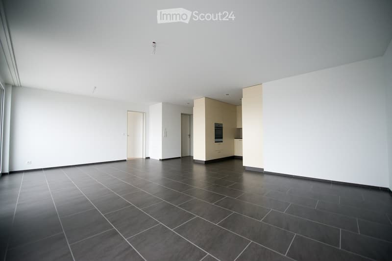 Empty room with dark tiled floor, white walls, and a small kitchen area with cabinets and an oven in the background.