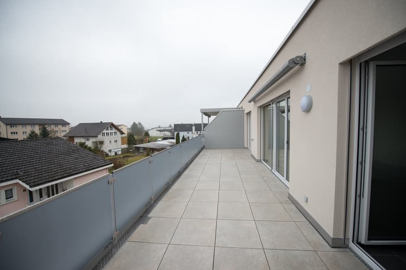 Balcony with tiled floor, glass sliding doors, and a low fence, overlooking a residential area with houses and trees.