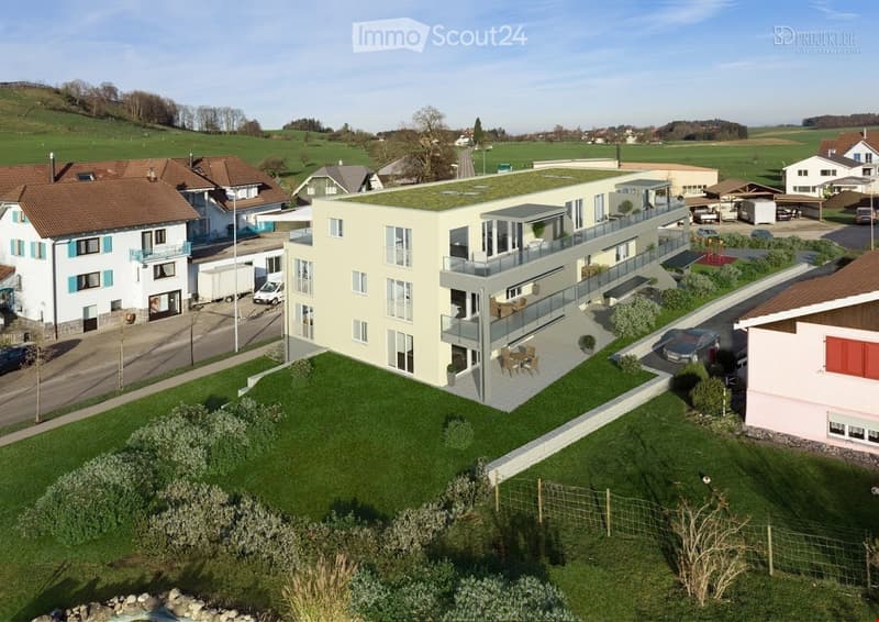 Modern multi-story building with a green roof, large windows, and a spacious balcony area. The surrounding area includes a well-maintained lawn, a parking lot, and neighboring houses.