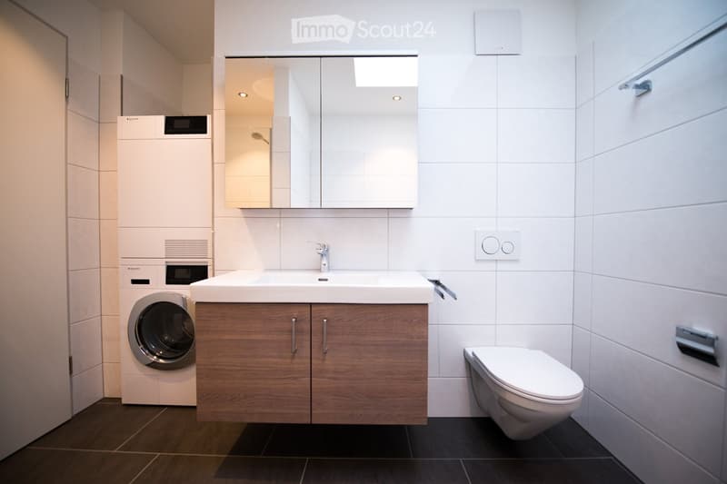 Modern bathroom with a white sink, mirror cabinet, toilet, and a private washing machine. The space features wooden cabinets and white tiled walls.