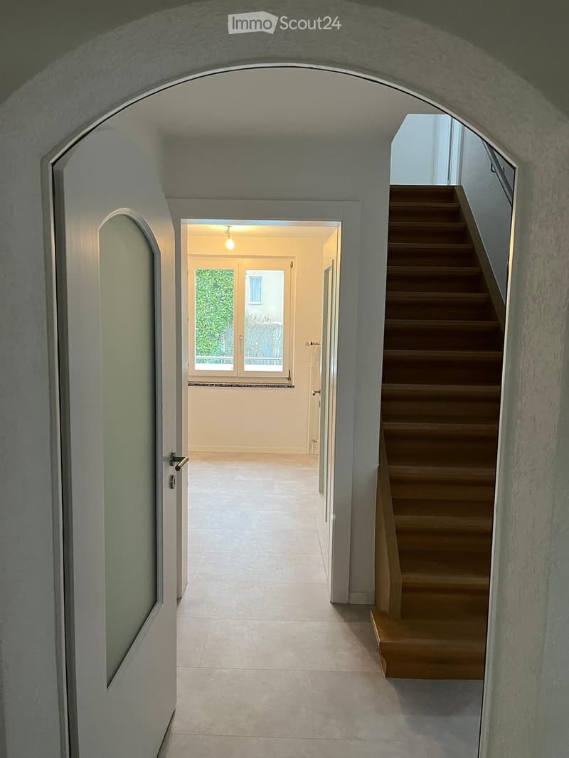 A modern hallway with a curved ceiling, tiled floor, and a wooden staircase leading upwards. A doorway opens to a brightly lit room with large windows and a view of greenery.