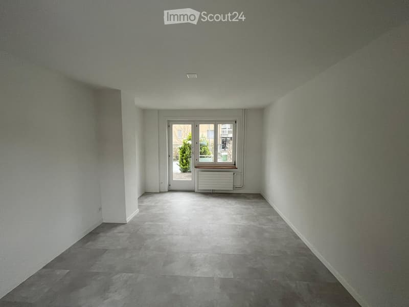 Empty room with light grey walls, grey tiled floor, and a large window with a radiator below it.