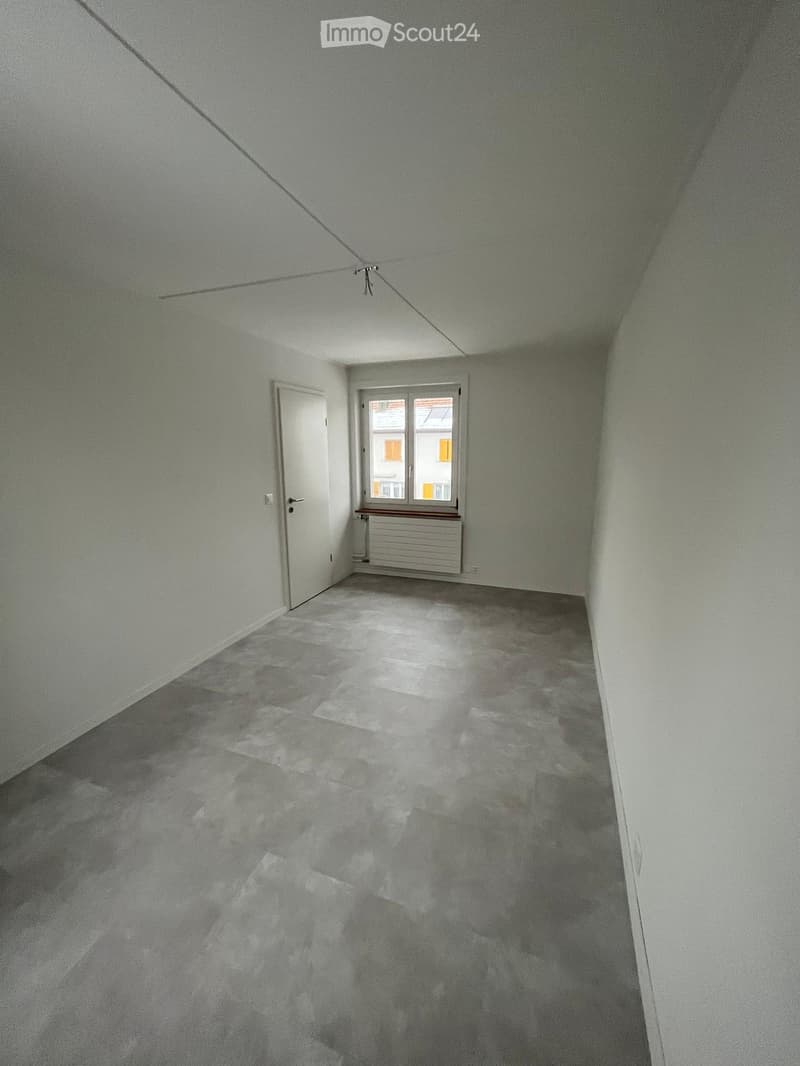 Empty room with light grey walls, a white door, a window with a radiator below, and a grey carpeted floor.