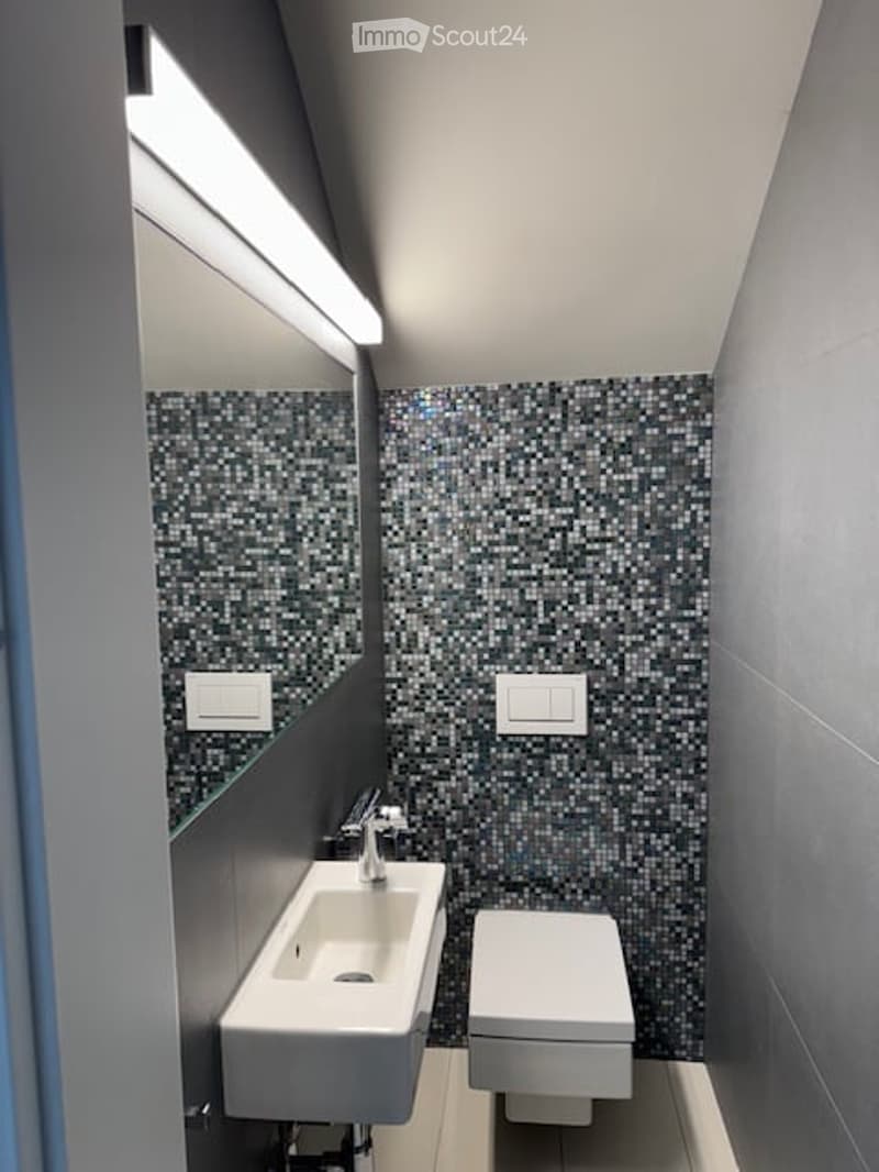 Modern, compact bathroom with a white sink, toilet, and a large mirror with built-in lighting. The walls are tiled in a mosaic pattern, and the space is well-lit with recessed lighting.