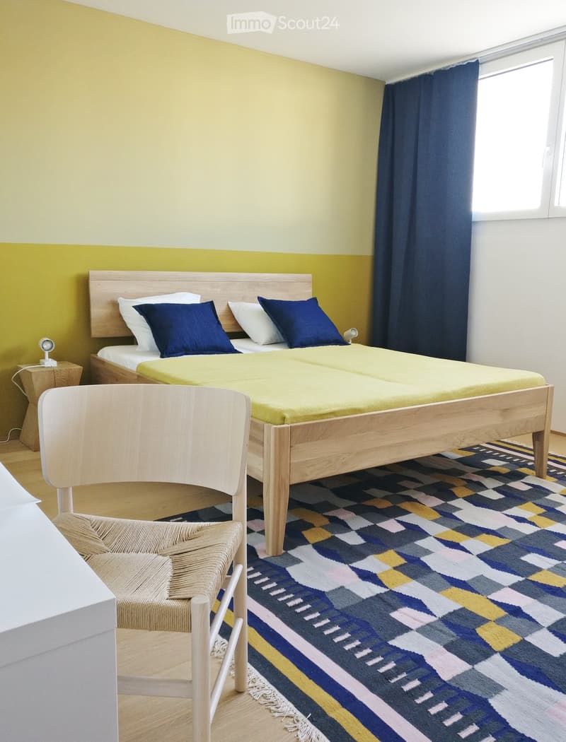 Bright bedroom with a double bed, two nightstands, a chair, and a colorful rug.