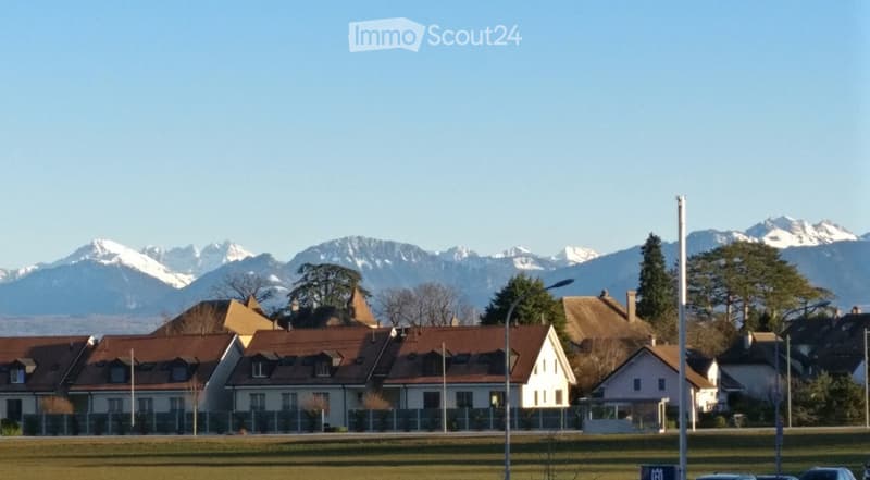 Residential houses with red-tiled roofs and white walls, surrounded by green fields and snow-capped mountains in the background.