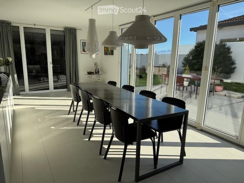 Bright dining area with a long black dining table surrounded by black chairs, pendant lights hanging from the ceiling, large glass sliding doors leading to an outdoor patio, and a small side table with a decorative item.