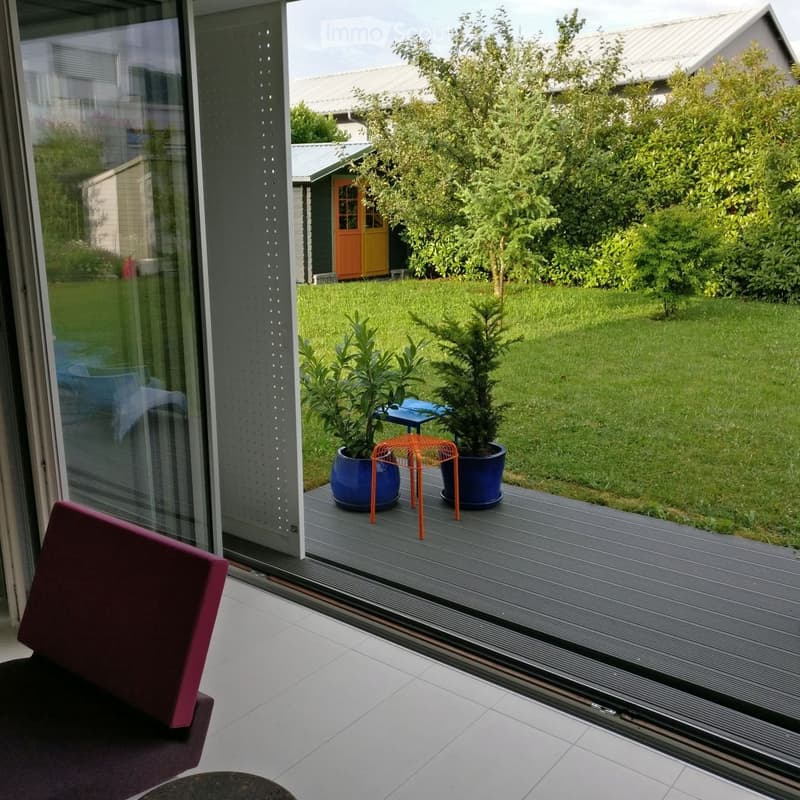 Balcony with a grey deck, two potted plants in blue pots, and an orange wire stool. The view shows a well-maintained lawn, trees, and a small shed with a yellow door.