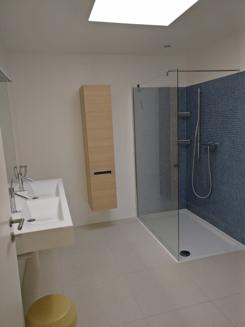 Modern bathroom with a white sink, wooden cabinet, glass-enclosed shower, and a yellow stool.
