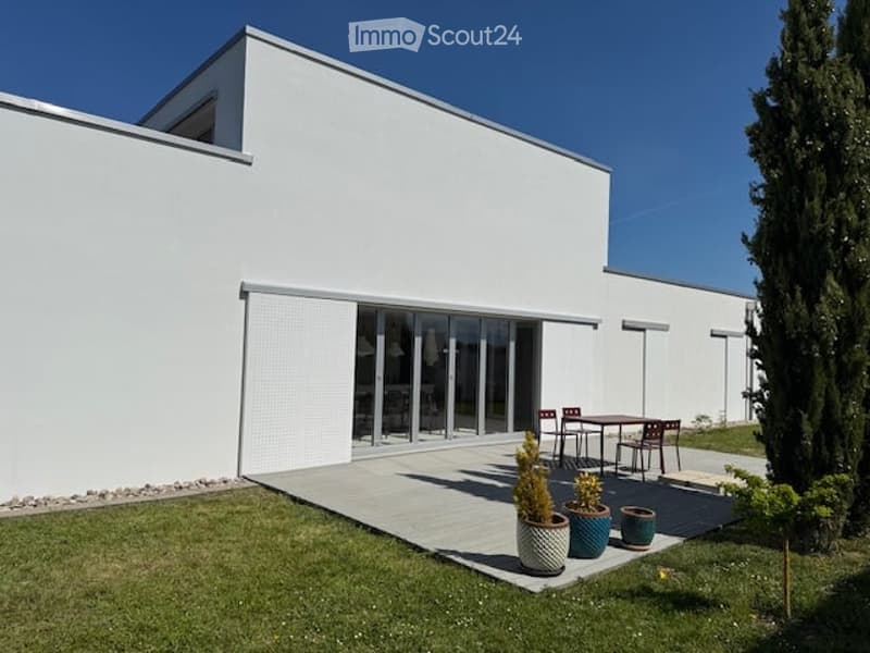 Modern white building with large glass sliding doors, outdoor patio with small table and chairs, potted plants, and a well-maintained lawn.