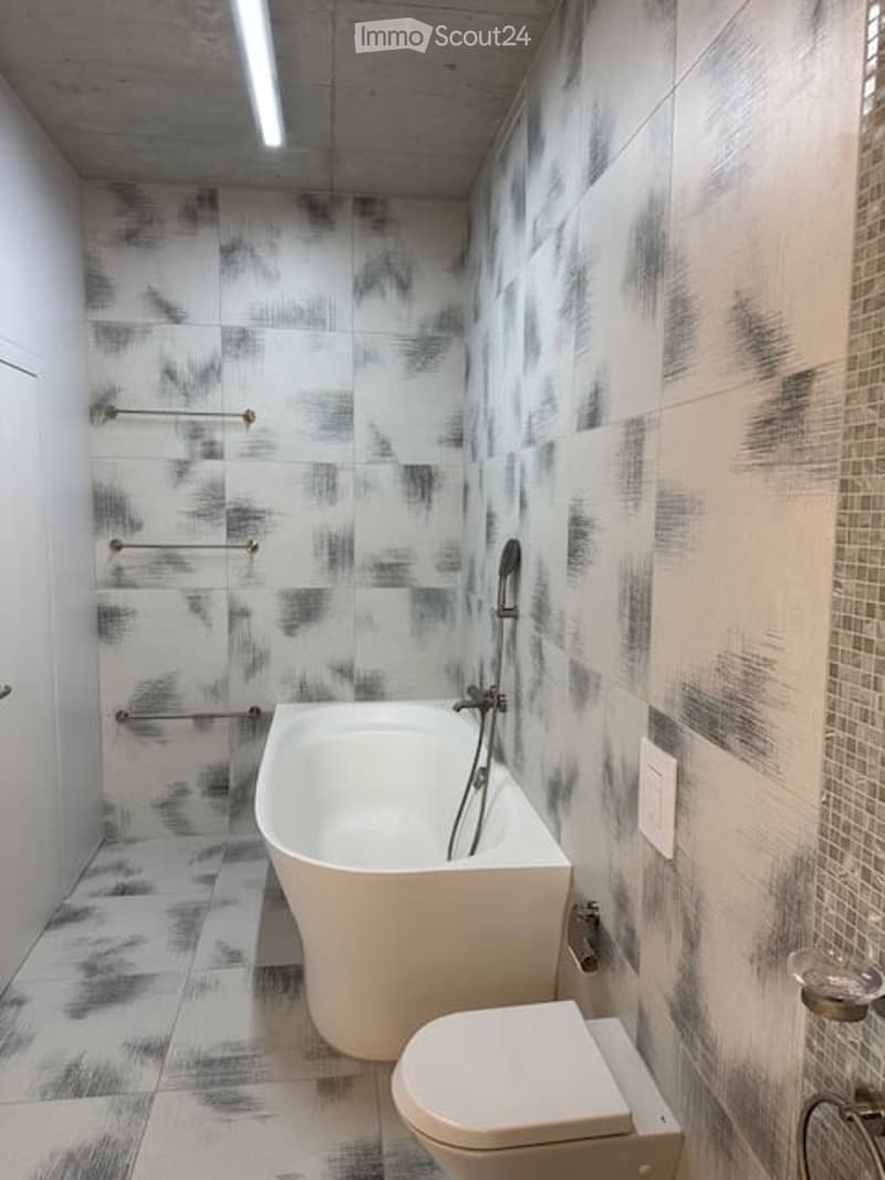 Modern bathroom with a freestanding bathtub, toilet, and a showerhead mounted on the wall. The walls are tiled with a black and white abstract pattern, and there are towel racks on the left side.