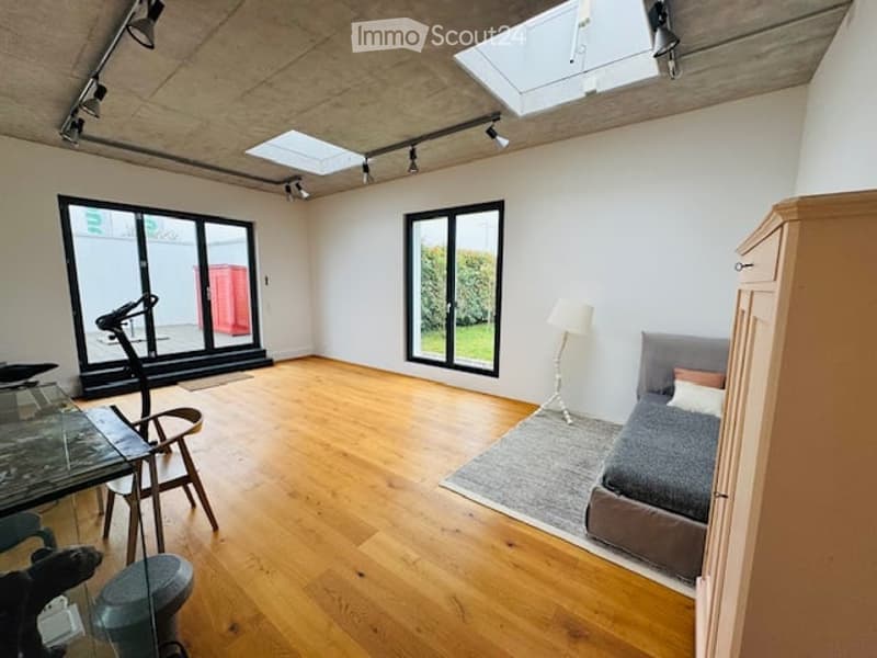 Bright, modern room with wooden flooring, large windows, a sofa, a table with chairs, a floor lamp, and an exercise bike.