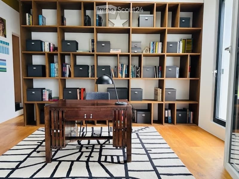 A modern, well-organized study or home office with a large wooden bookshelf filled with books and storage boxes, a wooden desk with a chair, and a sleek black desk lamp. The room features a geometric black-and-white rug on a light wooden floor.