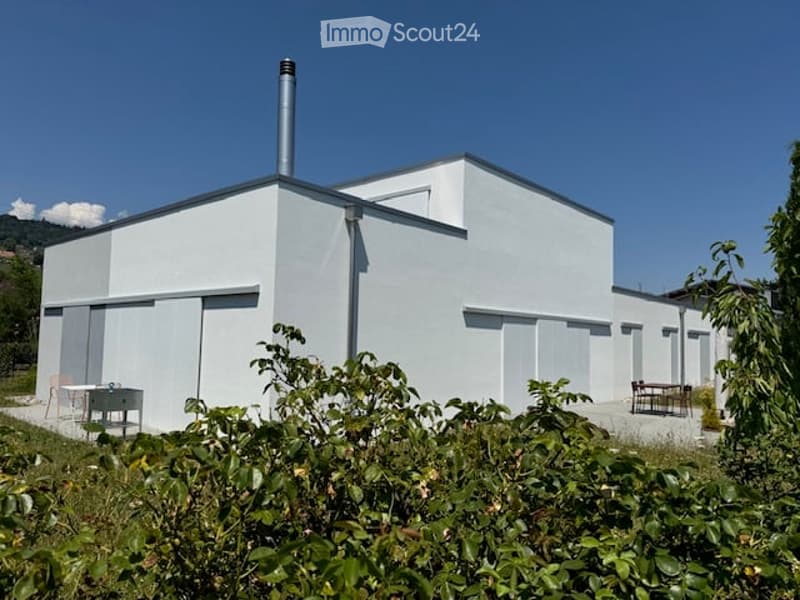 Modern, minimalist white building with a flat roof and chimney, surrounded by greenery and outdoor seating.