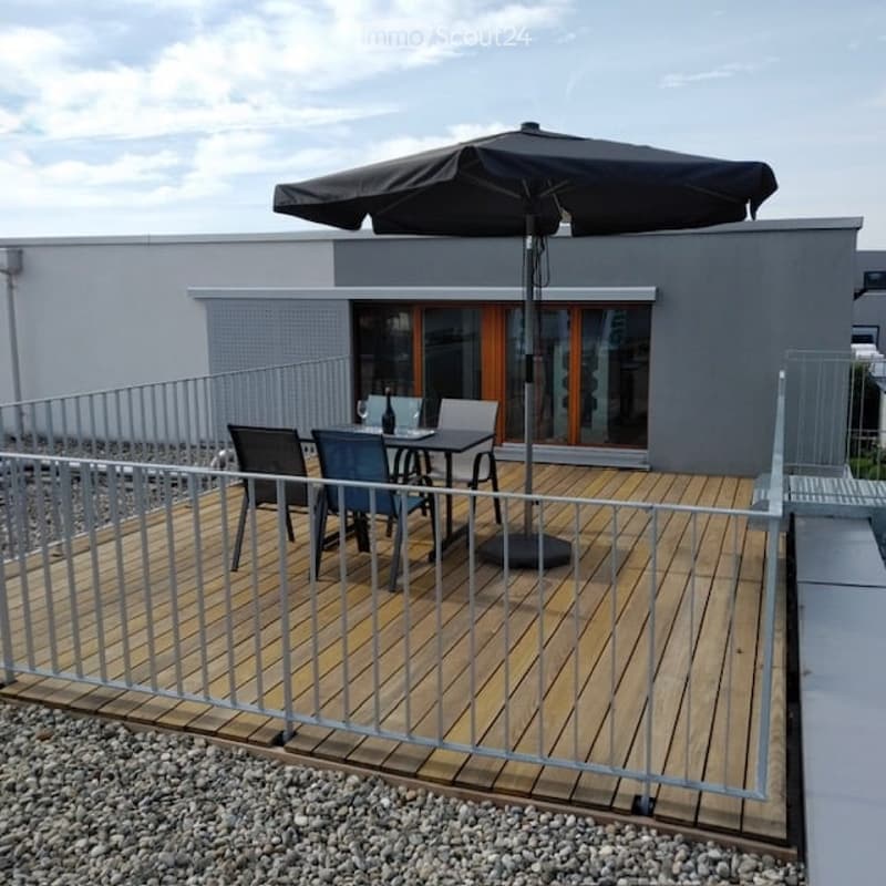 Wooden deck with metal railing, outdoor dining set, umbrella, and modern building in the background.