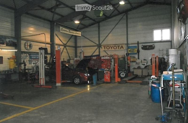 A spacious industrial workshop with a raised ceiling, exposed metal beams, and bright overhead lighting. A red Toyota car is on a lift, surrounded by various tools, equipment, and storage items. The walls feature signage, including a prominent "TOYOTA" lo