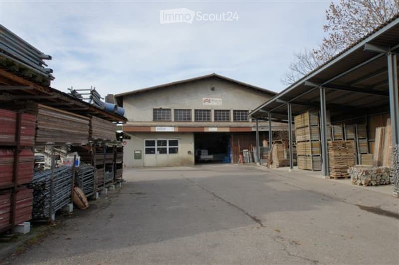 Warehouse building with a large open entrance, stacks of wooden planks and metal sheets on either side, and a sign above the entrance.