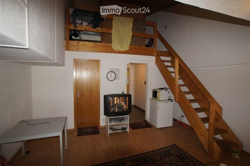 The room features a wooden floor, a staircase leading to a loft area, a TV on a stand, a small table, a refrigerator, and a microwave. The loft area has a bed and some clothing items.