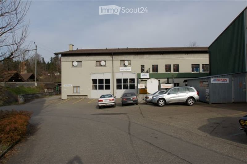 Single-story industrial or commercial building with a flat roof, large windows, and a parking area with several cars.