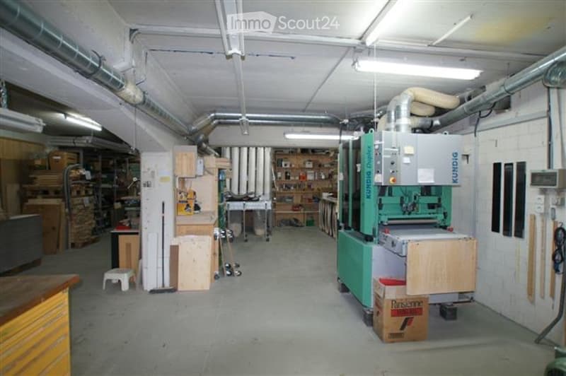 Workshop with industrial lighting, exposed pipes, various tools, and equipment including a saw and wooden planks.