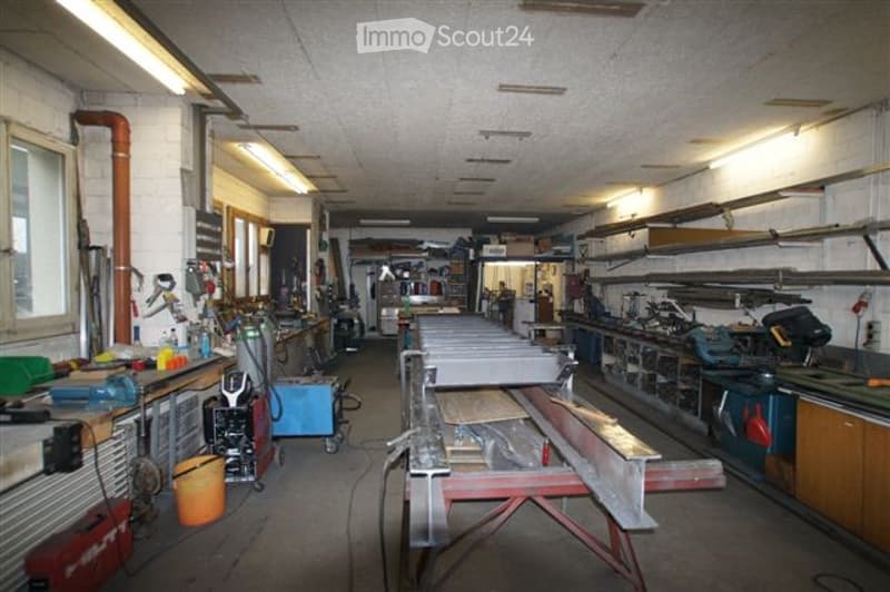 A spacious workshop with a concrete floor, exposed pipes, and ample lighting. The room features multiple workbenches, various tools, and storage cabinets.