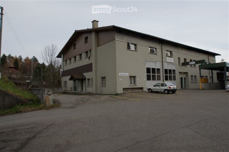 A single-story industrial or commercial building with a flat roof, multiple windows, and a small covered entrance. A white car is parked in the adjacent parking lot.
