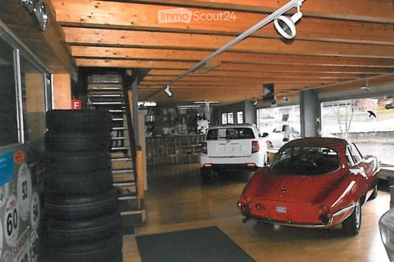 Garage with wooden ceiling and exposed beams, featuring a red sports car, white SUV, stacked tires, and shelves with items.